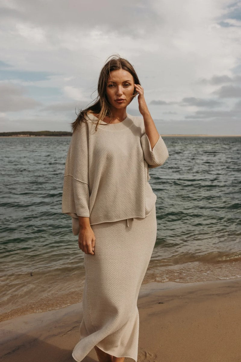 Woman in a beige outfit standing on a beach with ocean and cloudy sky in the background