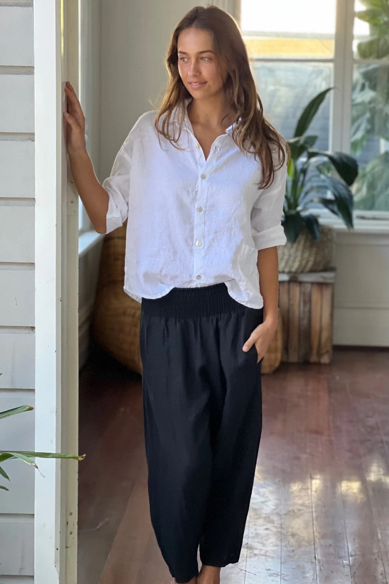 Woman wearing a white blouse and black pants standing in a room with wooden floor and plants.
