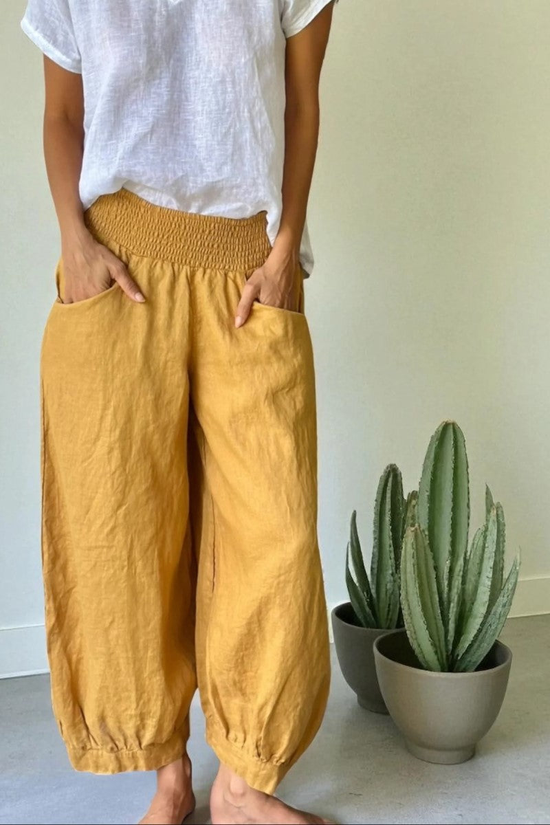 Person wearing mustard yellow pants and a white shirt standing next to two potted plants.