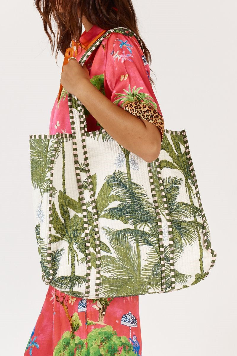 Person wearing a pink floral shirt and colorful pants, holding a green palm leaf patterned bag.