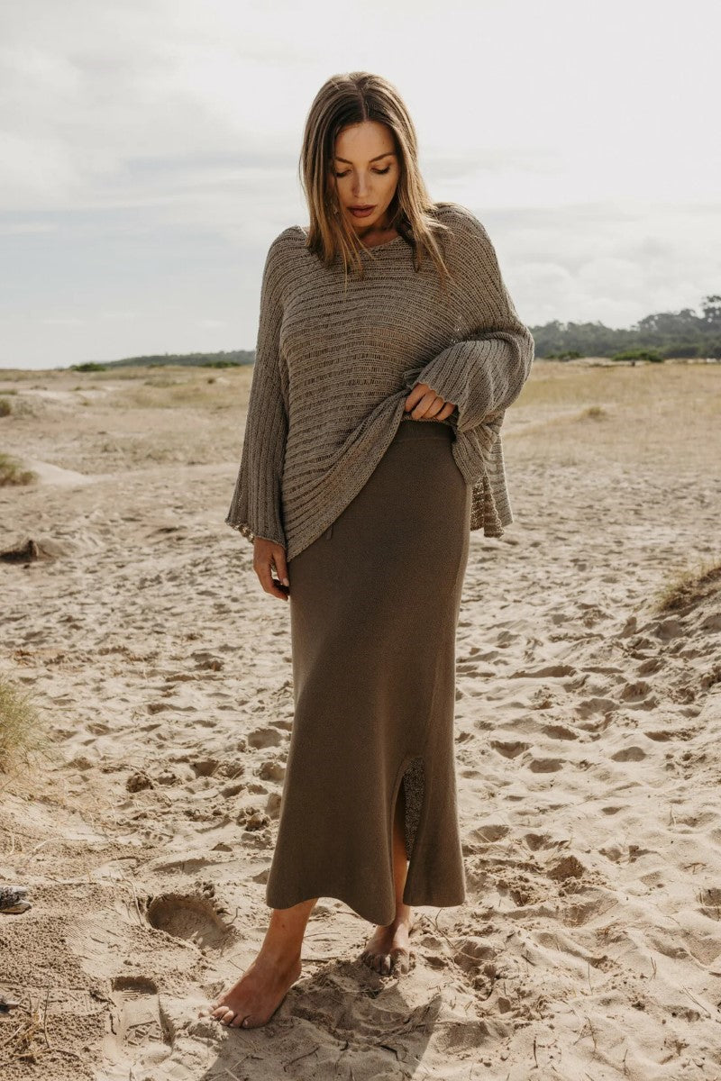 Woman wearing a brown sweater and skirt standing on a sandy beach.