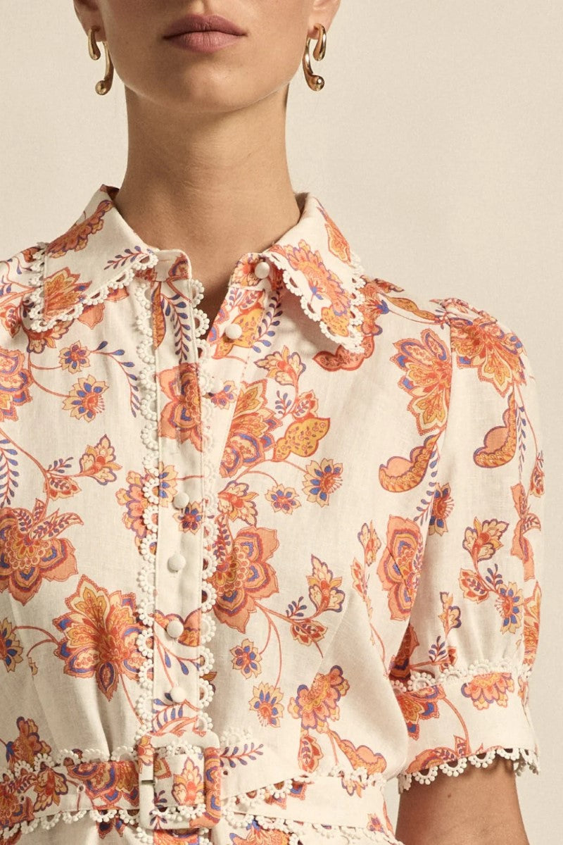 Close-up of a person wearing a floral blouse with orange and purple patterns on a beige background.