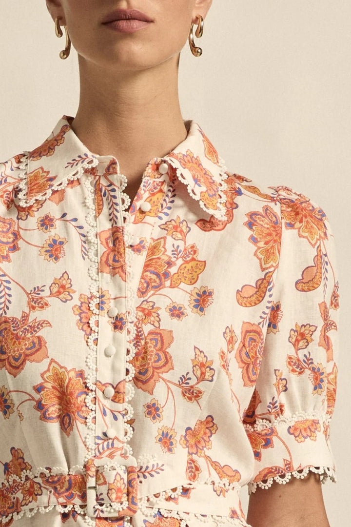 Close-up of a person wearing a floral blouse with orange and purple patterns on a beige background.
