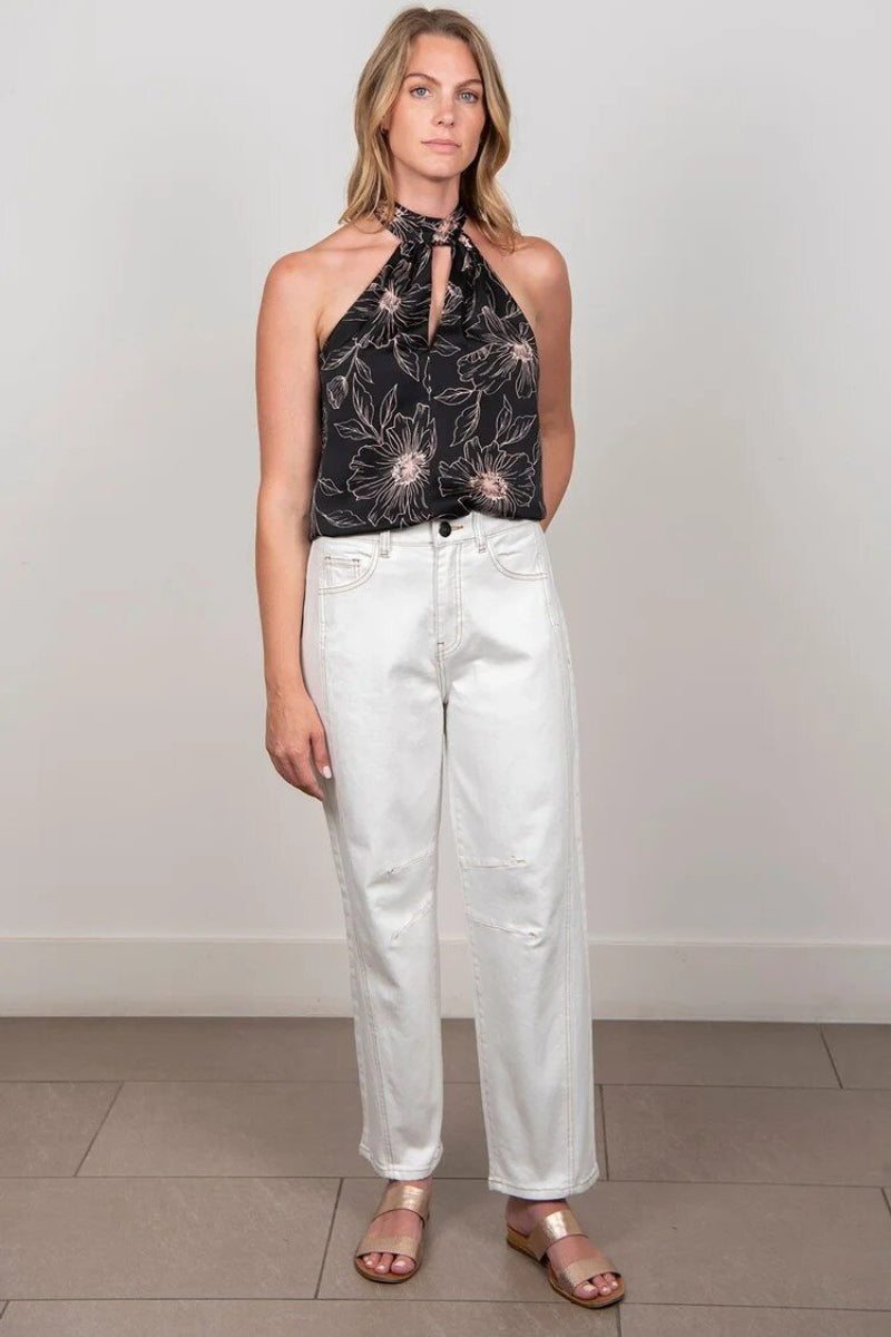 A woman modeling a black floral halter top paired with white high-waisted wide-leg jeans and metallic sandals, standing against a plain light gray wall.