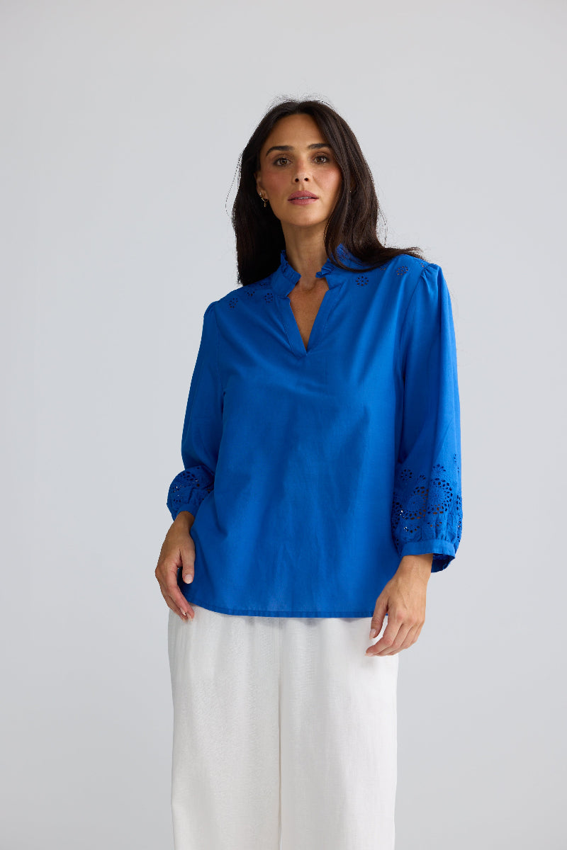 A woman modeling a vibrant royal blue long-sleeve blouse with lace detailing on the cuffs, paired with white trousers, against a solid light gray background.