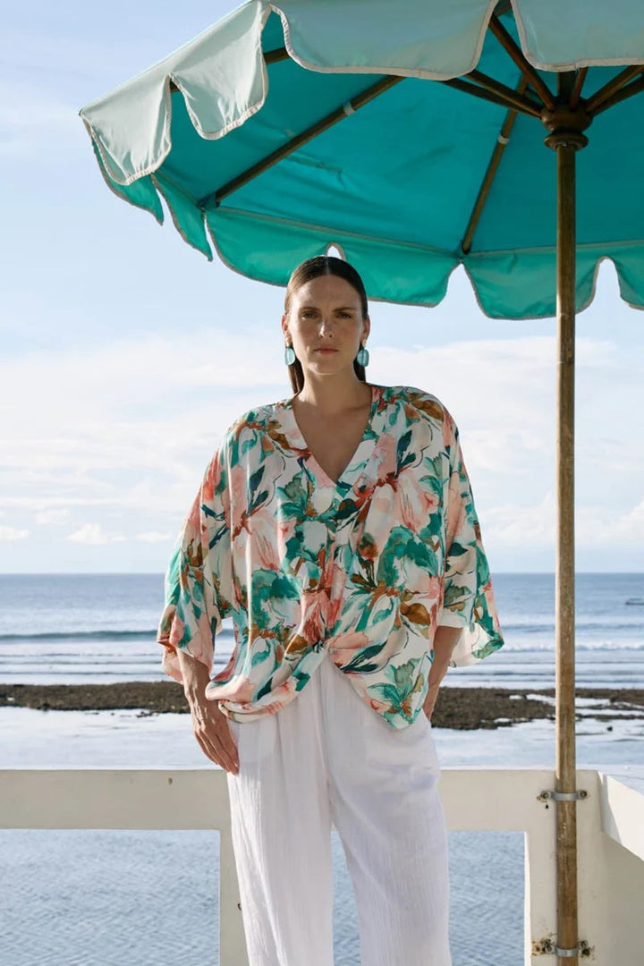 Woman in a floral blouse and white pants standing under a teal umbrella by the ocean.