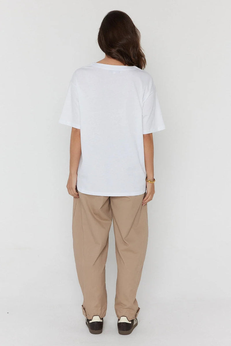 A woman viewed from behind wearing an oversized white short-sleeve t-shirt, loose beige pants, and dark loafers against a plain white studio background.