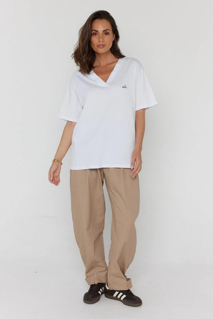 A woman modeling an oversized white short-sleeve V-neck T-shirt with a small embroidered logo on the chest, paired with loose beige pants and black sneakers, against a plain light gray studio background.