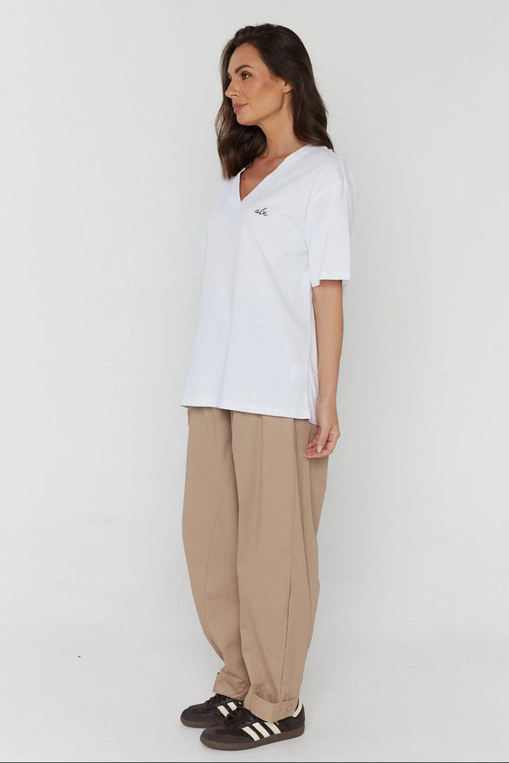 A woman modeling a white short-sleeve V-neck t-shirt with a small black embroidered logo on the left chest, paired with beige wide-leg trousers and black sneakers with white stripes, set against a plain white studio background.
