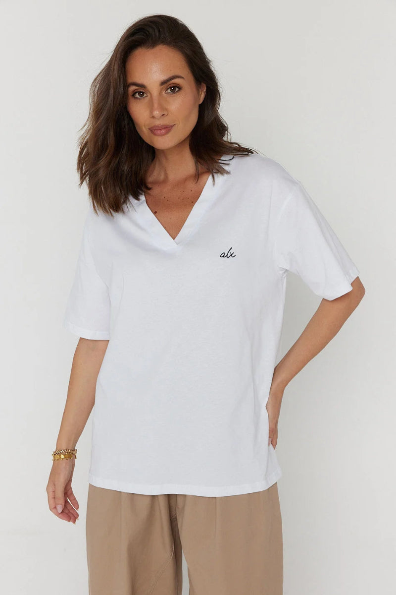 A woman modeling a white short-sleeve V-neck top with a small black script logo on the chest, paired with beige trousers, against a plain light gray background.