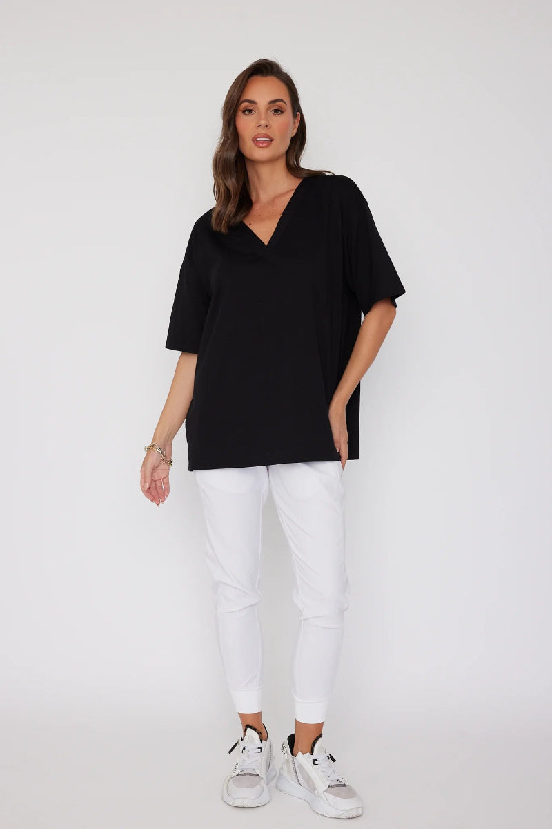 A woman modeling an oversized black short-sleeve V-neck top paired with white pants and white sneakers, standing against a plain white studio background.