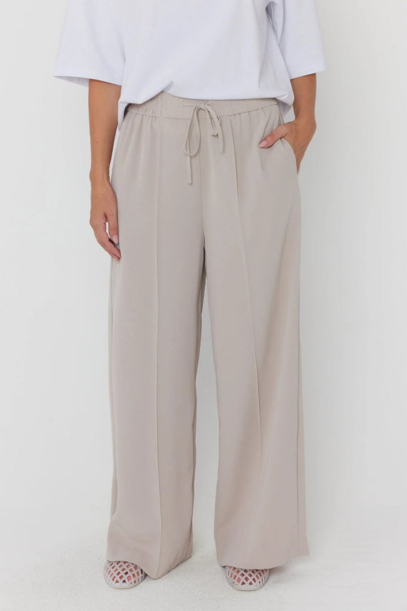 A person wearing loose, wide-leg drawstring trousers in a light warm gray-beige tone, paired with a white top and patterned shoes, photographed against a plain light background.