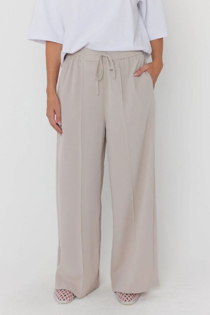 A person wearing loose, wide-leg drawstring trousers in a light warm gray-beige tone, paired with a white top and patterned shoes, photographed against a plain light background.