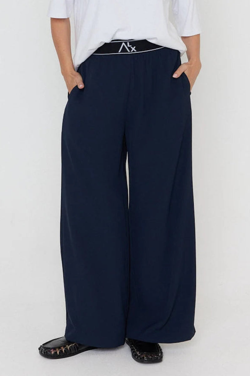 A person wearing dark navy wide-leg trousers with a black elastic waistband featuring a white logo, paired with a white t-shirt and black shoes, against a plain white background.