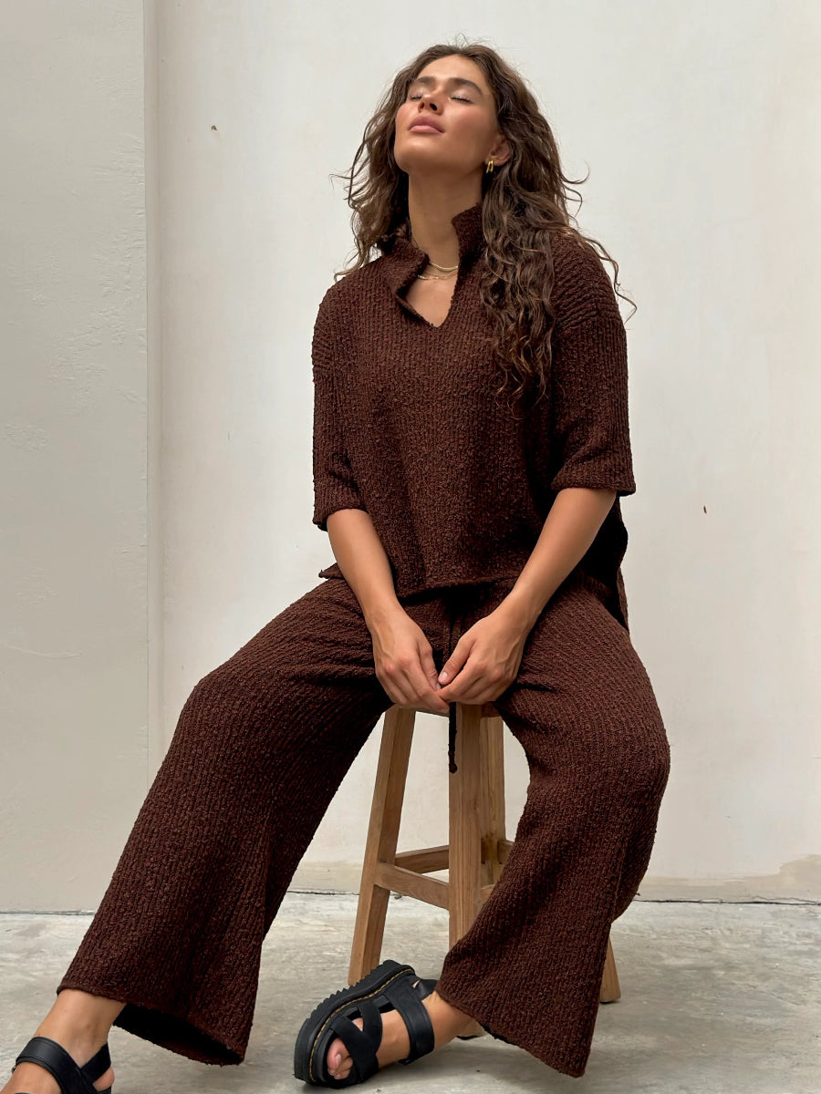 Woman sitting on stool wearing brown knit lounge set and sandals.