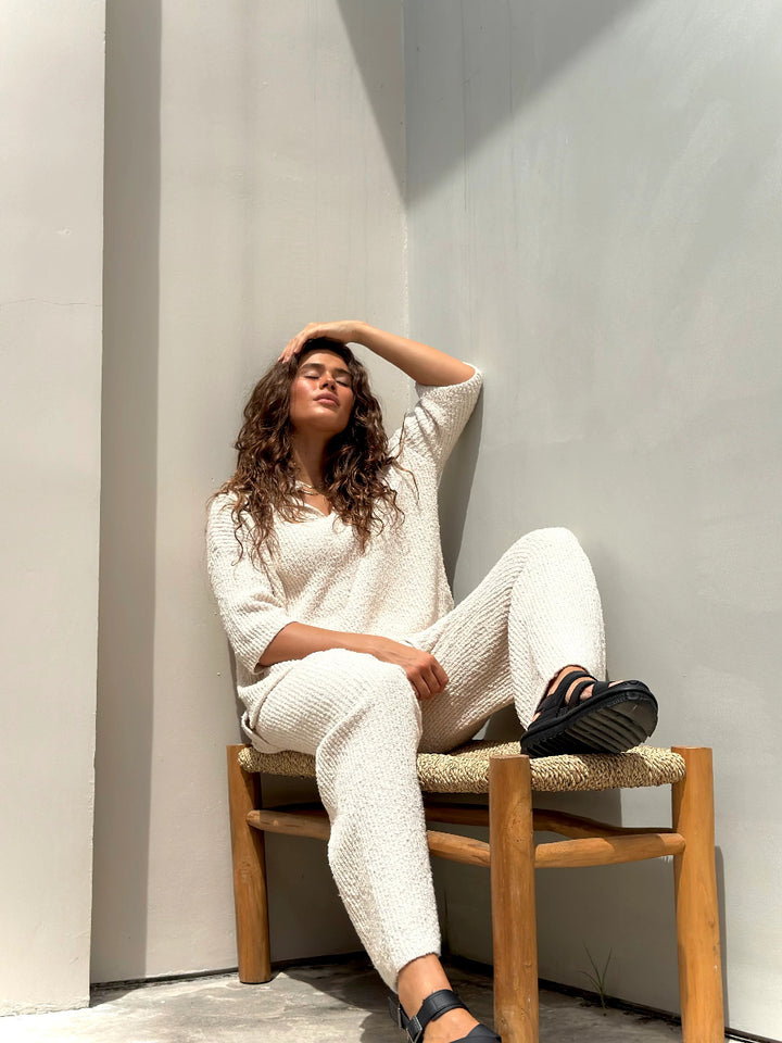 Woman seated in cream knit lounge set on woven bench.