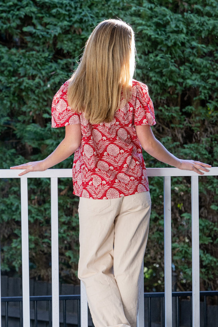 Woman wearing a red, floral patterned blouse with flared sleeves standing outdoors with greenery in the background