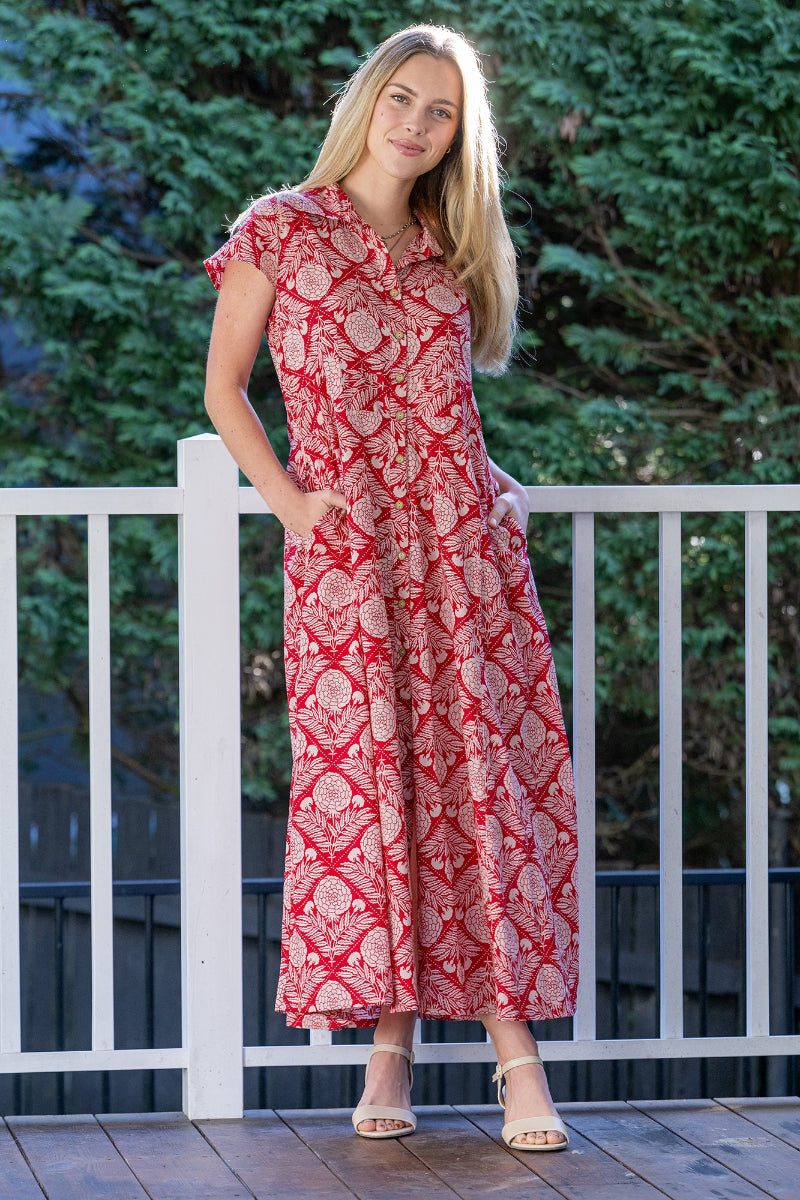 Woman wearing a red, floral patterned maxi dress standing on a wooden deck with greenery in the background.