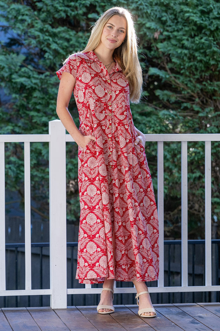 Woman wearing a red, floral patterned maxi dress standing on a wooden deck with greenery in the background.