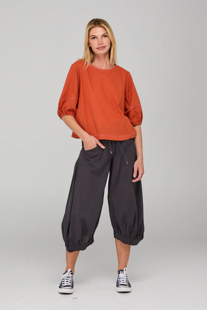 Woman wearing a rust-colored top and dark pants on a plain background
