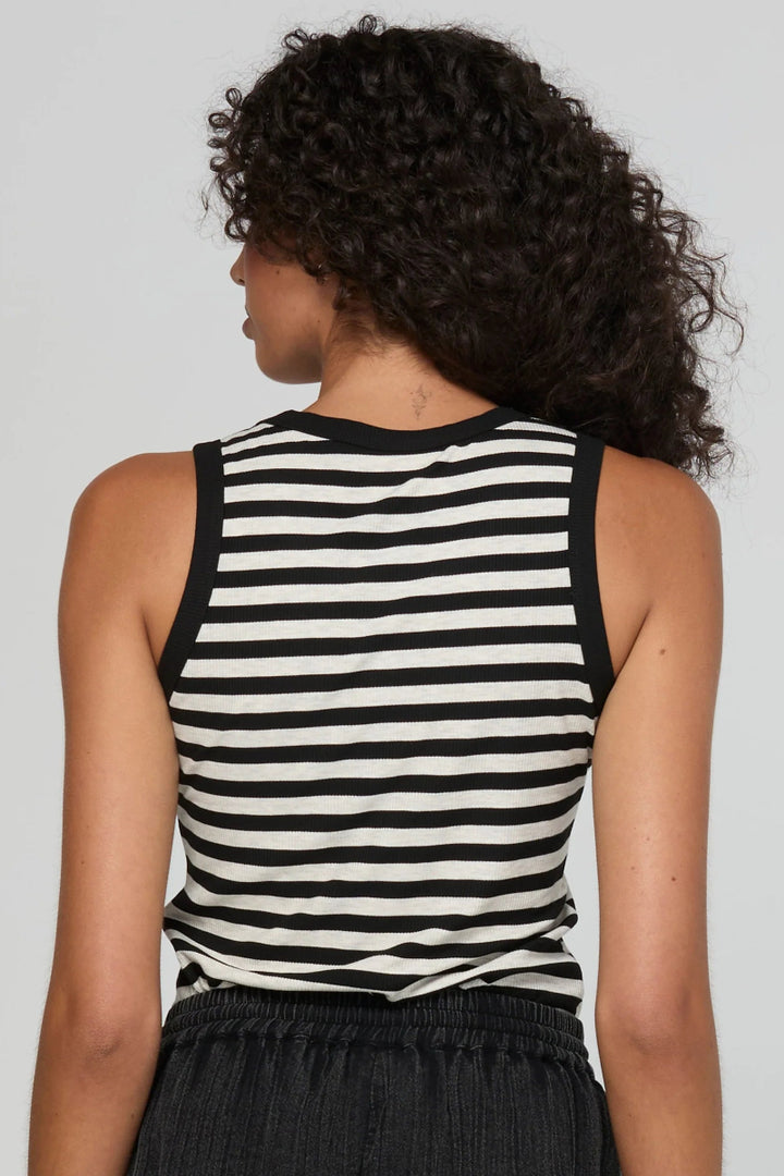 A woman shown from the back wearing a black and white horizontally striped sleeveless tank top and dark gray textured pants, set against a solid light gray studio background.