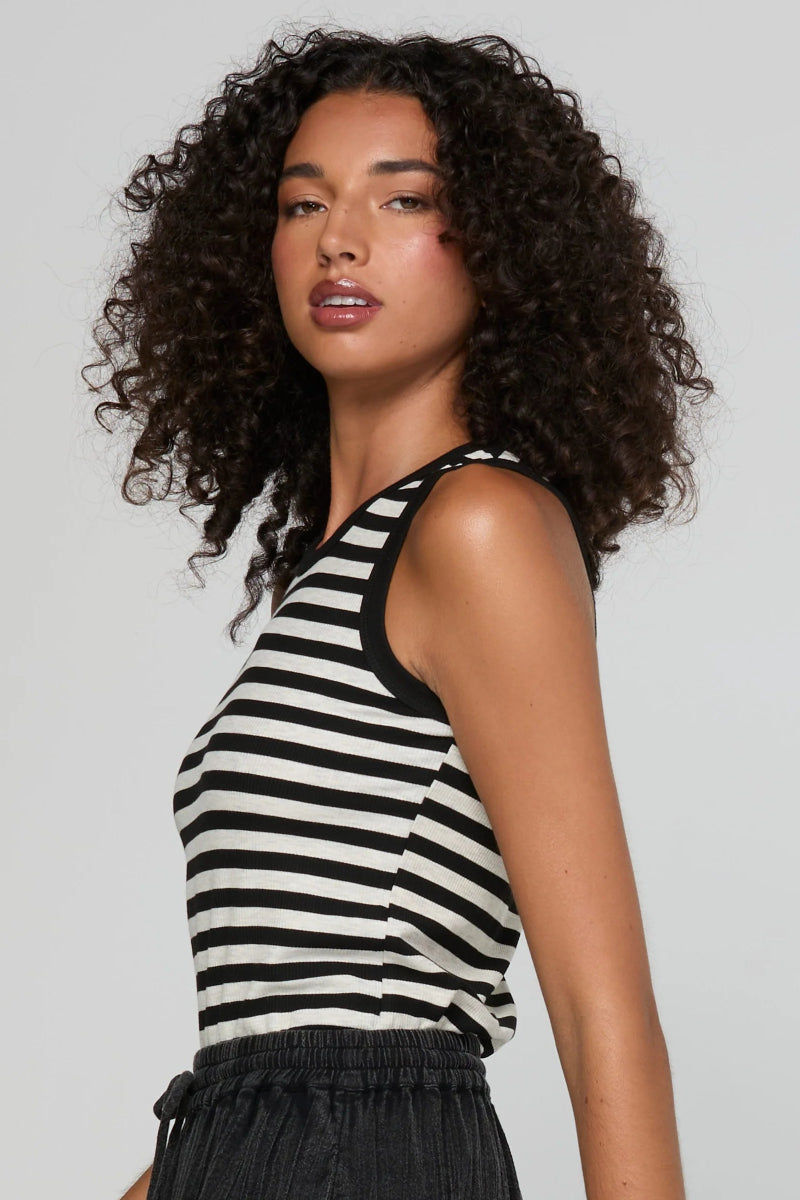 A woman with curly dark hair wearing a black and white horizontally striped sleeveless tank top, shown from a side-back angle against a light gray studio background.