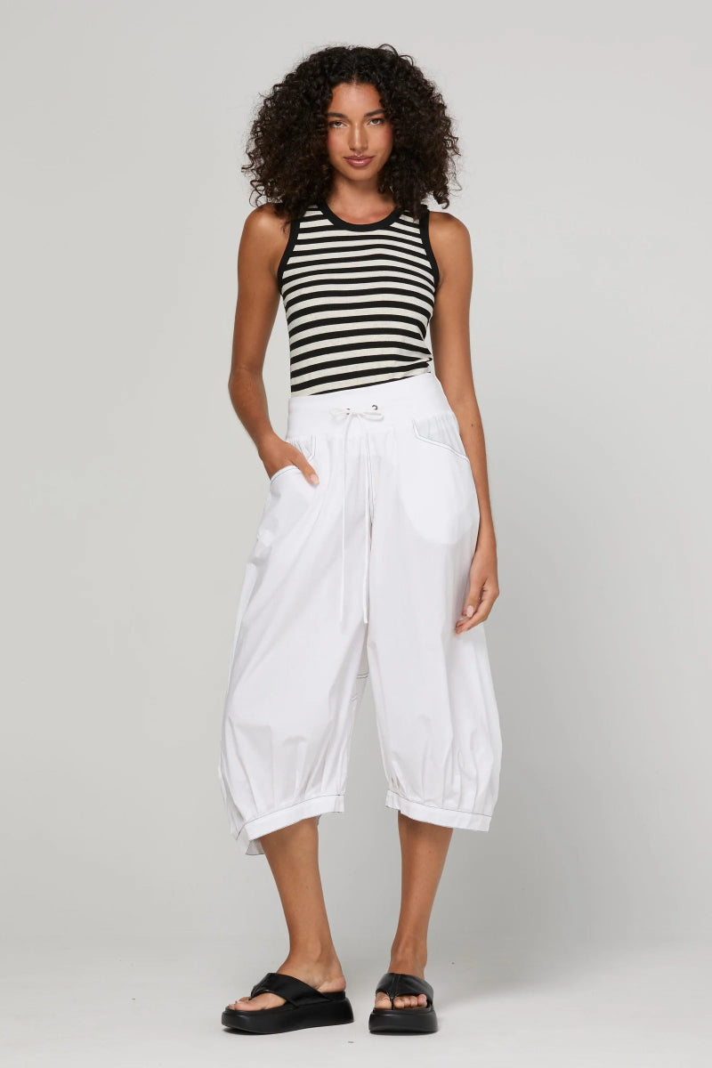 A model wearing a black and white striped sleeveless crop top, white high-waisted cropped pants, and black slide sandals, standing against a light gray studio background.