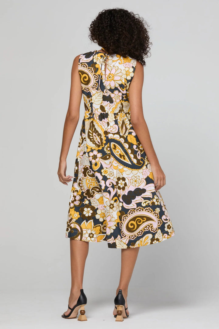 A woman viewed from the back wearing a sleeveless knee-length dress with a bold floral and paisley print in black, yellow, white, and pink, paired with black heeled sandals against a light gray studio background.