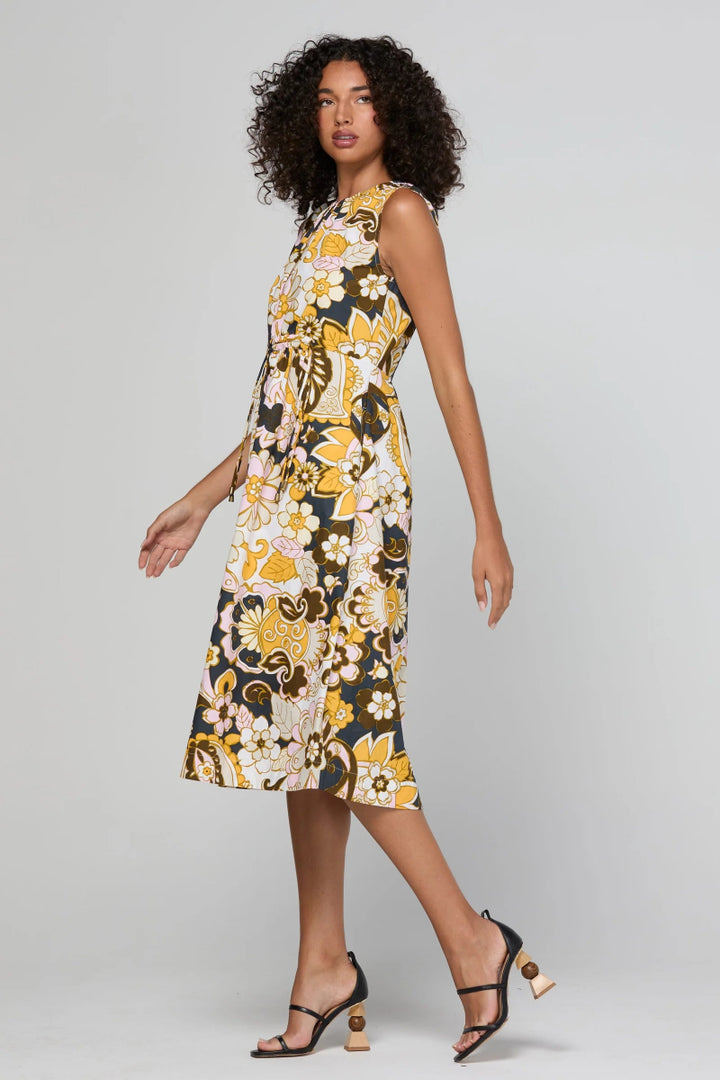 A woman modeling a sleeveless midi dress with a bold yellow, white, and black floral pattern, paired with black strappy heels, against a light gray studio background.