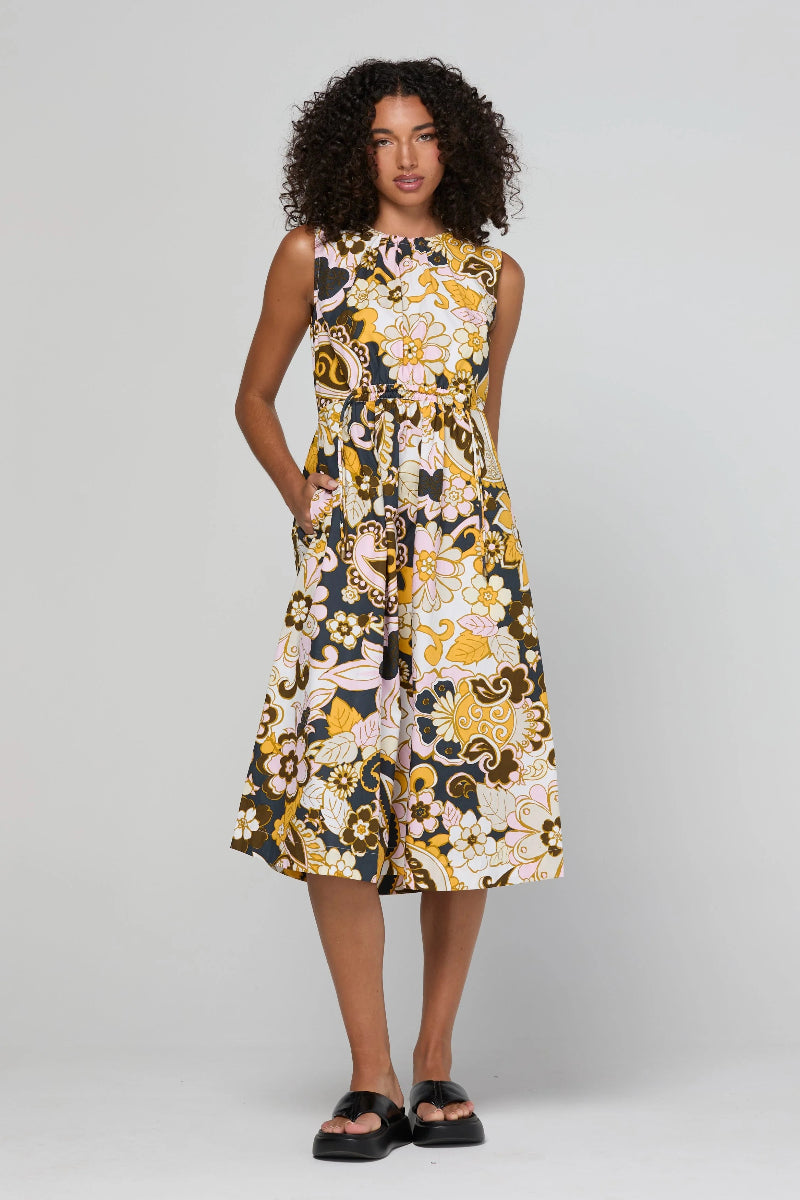 A woman modeling a sleeveless midi dress with a bold floral and abstract print in yellow, black, white, and beige, paired with black slide sandals, against a light gray studio background.