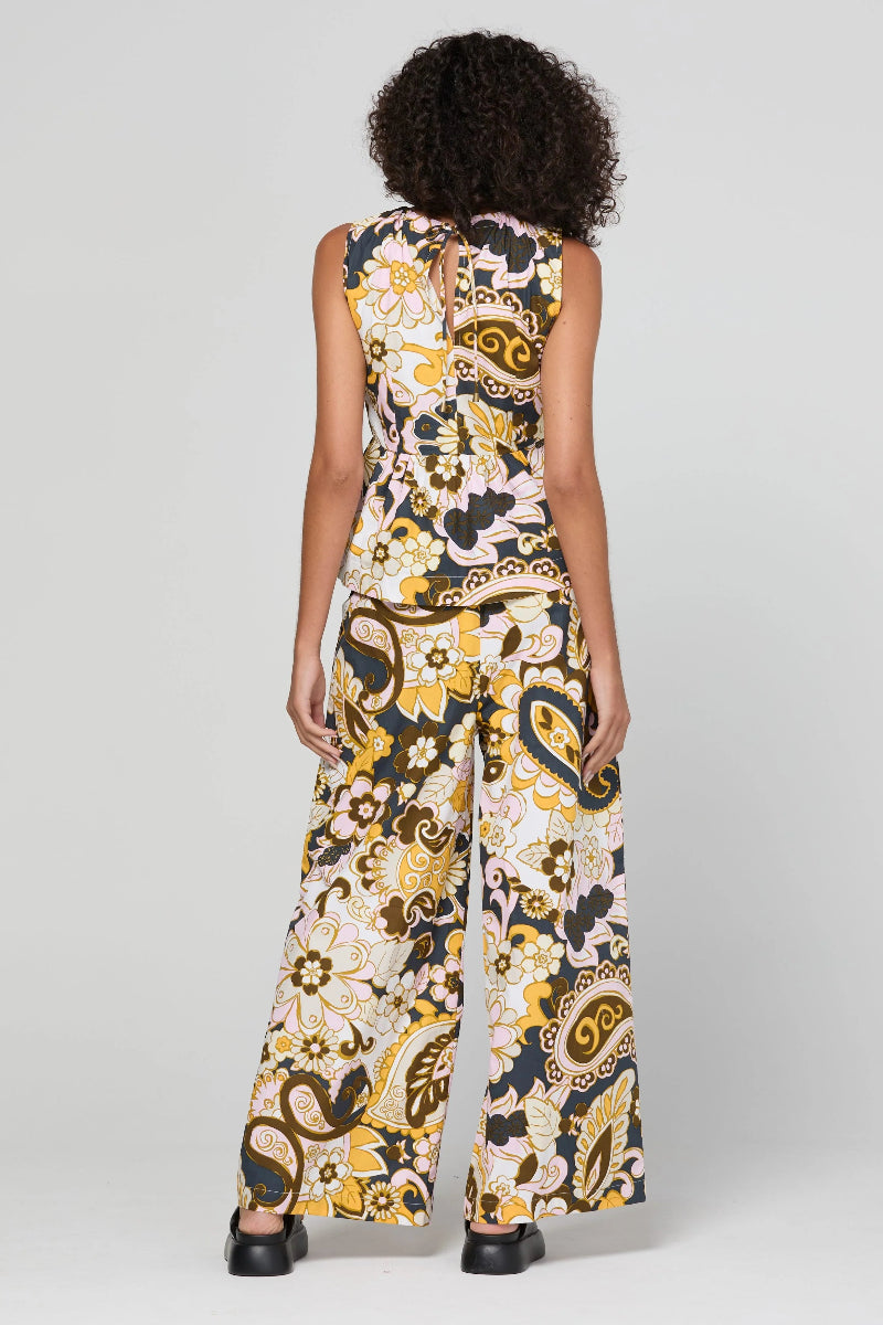 A model viewed from the back wearing a sleeveless wide-leg jumpsuit with a bold yellow, black, and white paisley pattern, set against a plain light gray studio background.