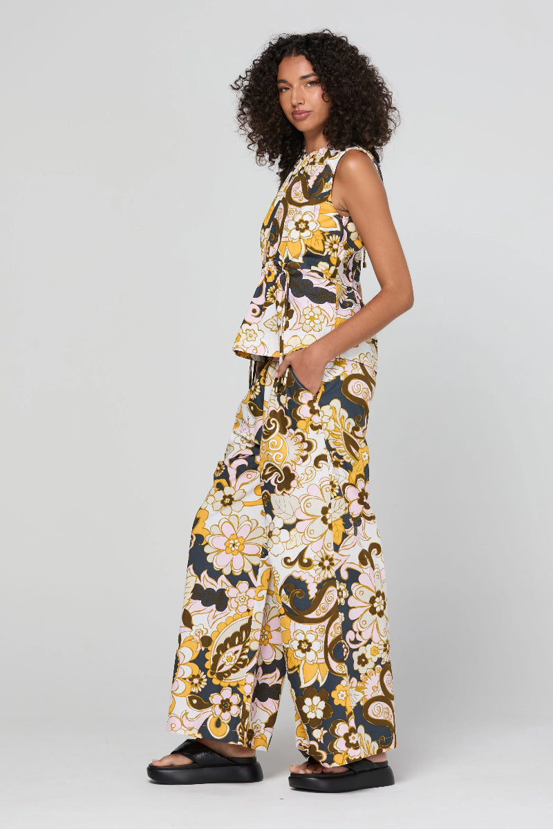 A model wearing a sleeveless matching set of a peplum top and wide-leg trousers in a yellow, black, and white floral paisley print, standing against a light gray studio background.