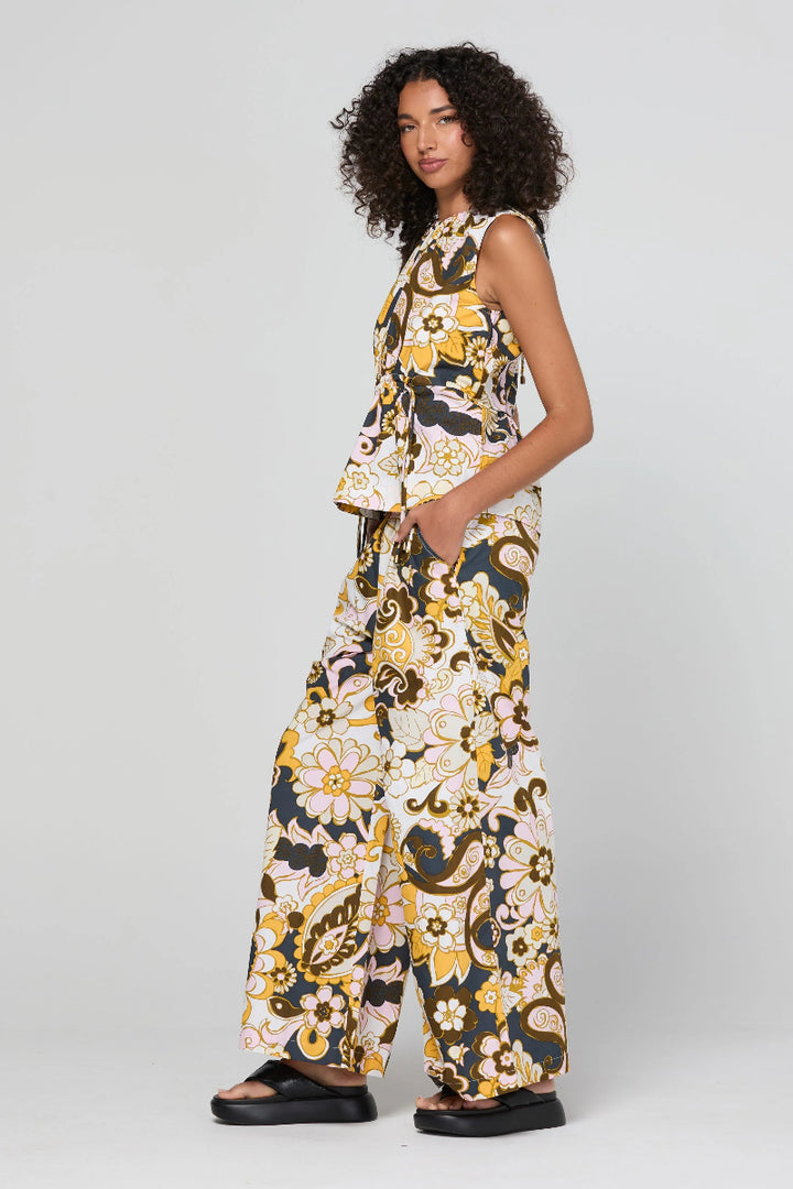A model wearing a sleeveless matching set of a peplum top and wide-leg trousers in a yellow, black, and white floral paisley print, standing against a light gray studio background.