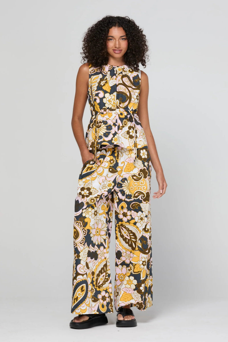 A woman modeling a sleeveless jumpsuit with a wide-leg pant leg and a high gathered neckline. The jumpsuit features a bold, multicolor paisley and floral pattern in yellow, white, black, and brown.