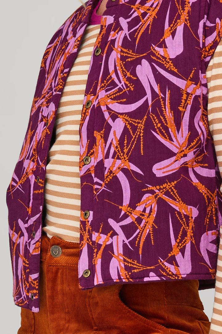 Person wearing a purple patterned vest over a striped shirt and brown pants on a light gray background