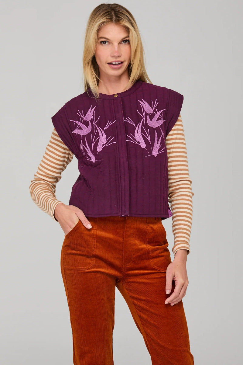 Woman wearing a purple vest with floral patterns, striped shirt, and brown pants on a gray background