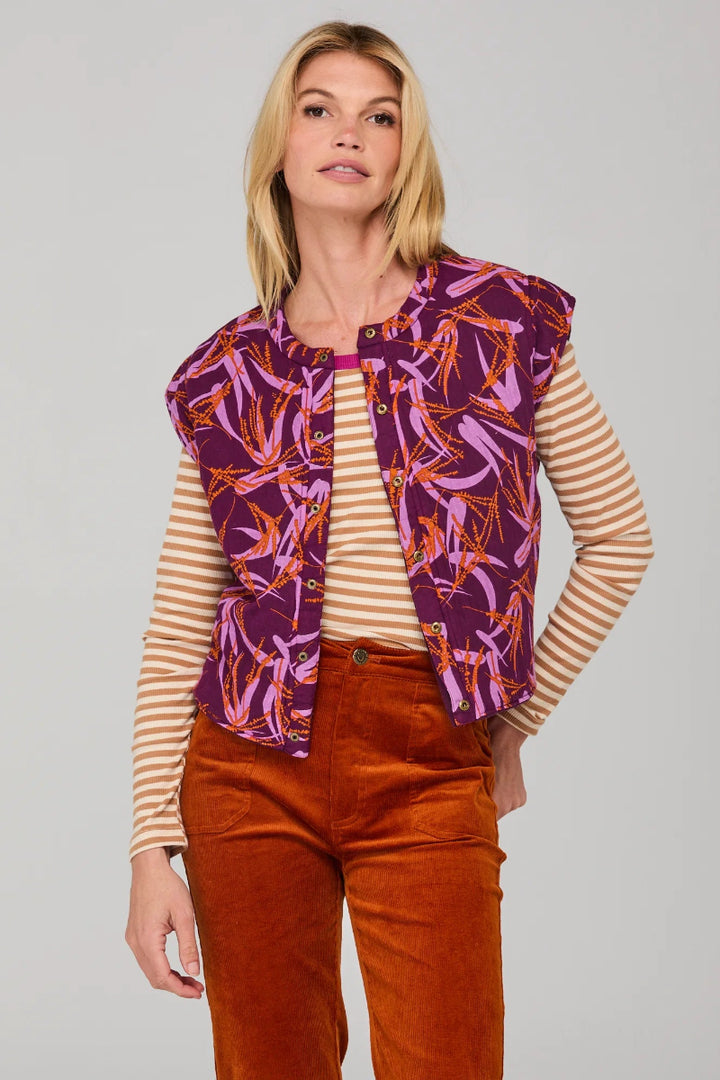 Woman wearing a patterned purple vest over a striped shirt with brown pants on a gray background