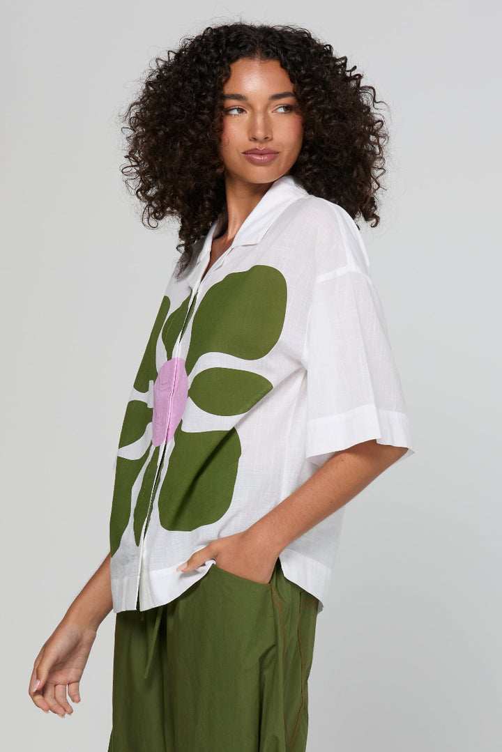 Woman wearing a white shirt with a large green floral design and green pants on a light gray background