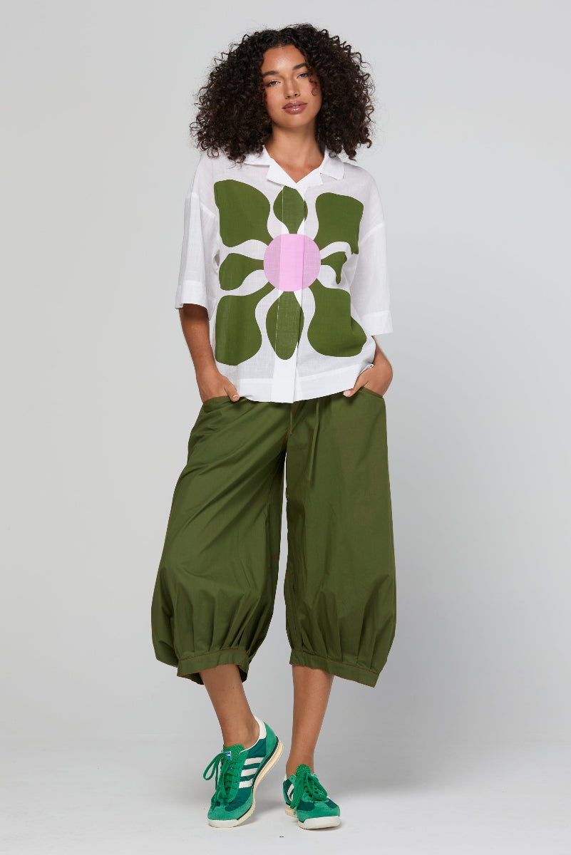 Woman wearing a white shirt with a large green floral design and green pants on a plain background