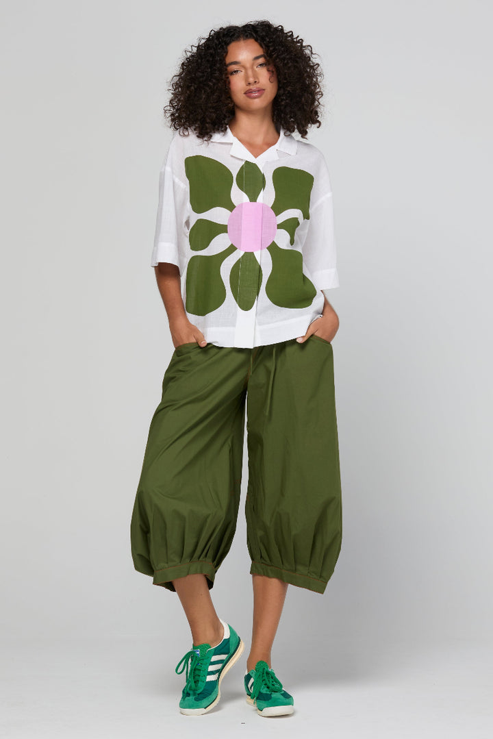 Woman wearing a white shirt with a large green floral design and green pants on a plain background