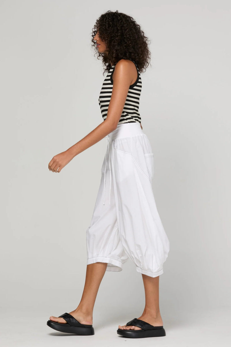 A model is shown from a side profile wearing white balloon-style cropped pants, a black and white striped tank top, and black slide sandals against a light gray studio background.