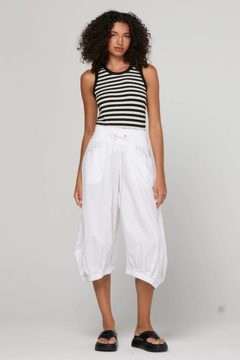 A model stands against a light gray studio background wearing a black and white horizontally striped sleeveless top, loose white cropped pants, and black slide sandals.