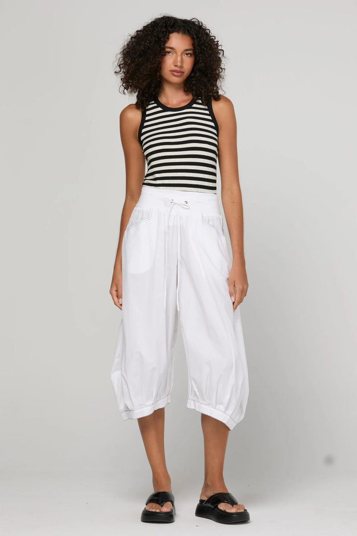 A model stands against a light gray studio background wearing a black and white horizontally striped sleeveless top, loose white cropped pants, and black slide sandals.