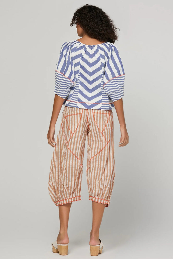 A woman shown from the back wearing a blue and white chevron-patterned top with ruffled sleeves and striped cropped pants in beige and orange, standing against a light gray studio background.