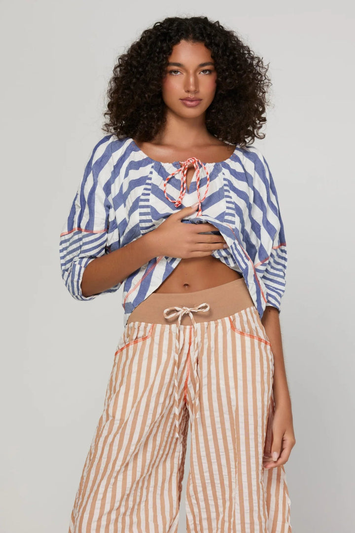 A model wearing a blue and white striped crop top with orange tie details and beige and white striped wide-leg pants, photographed against a neutral light gray background.