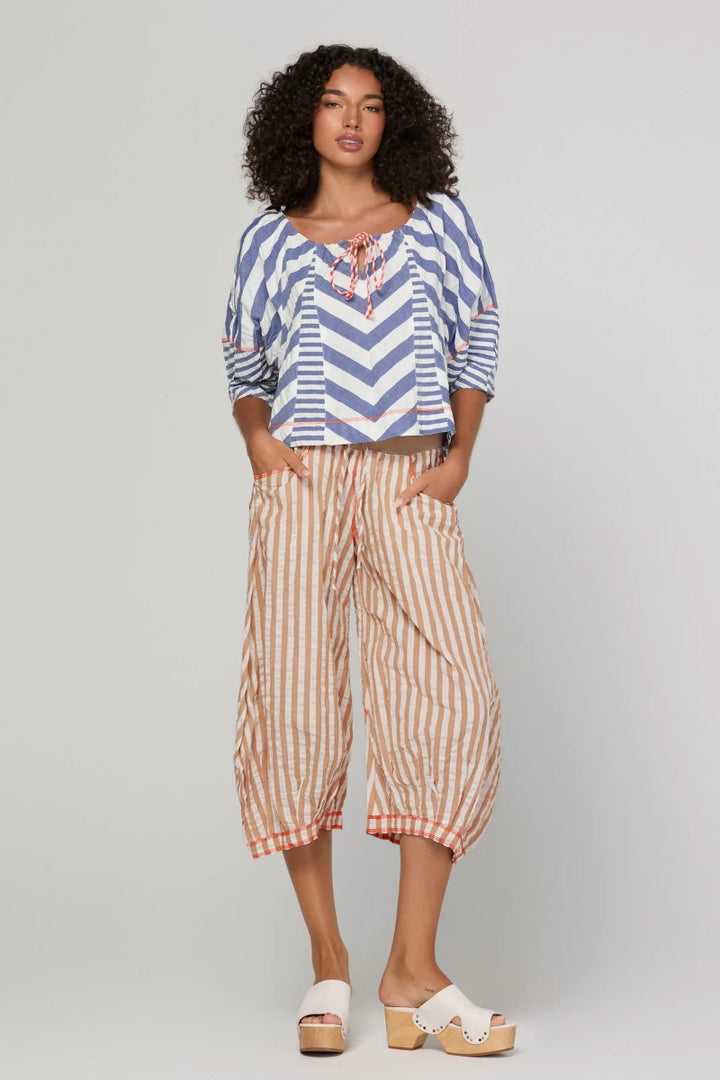 A woman modeling a two-piece outfit consisting of a blue and white chevron-patterned top with orange tie details and beige-and-orange vertically striped cropped pants, paired with white slide sandals, against a light gray studio background.