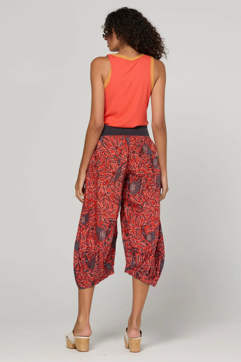 A woman shown from the back wearing a sleeveless coral-orange top and red-and-black patterned cropped pants, photographed against a light gray studio background.