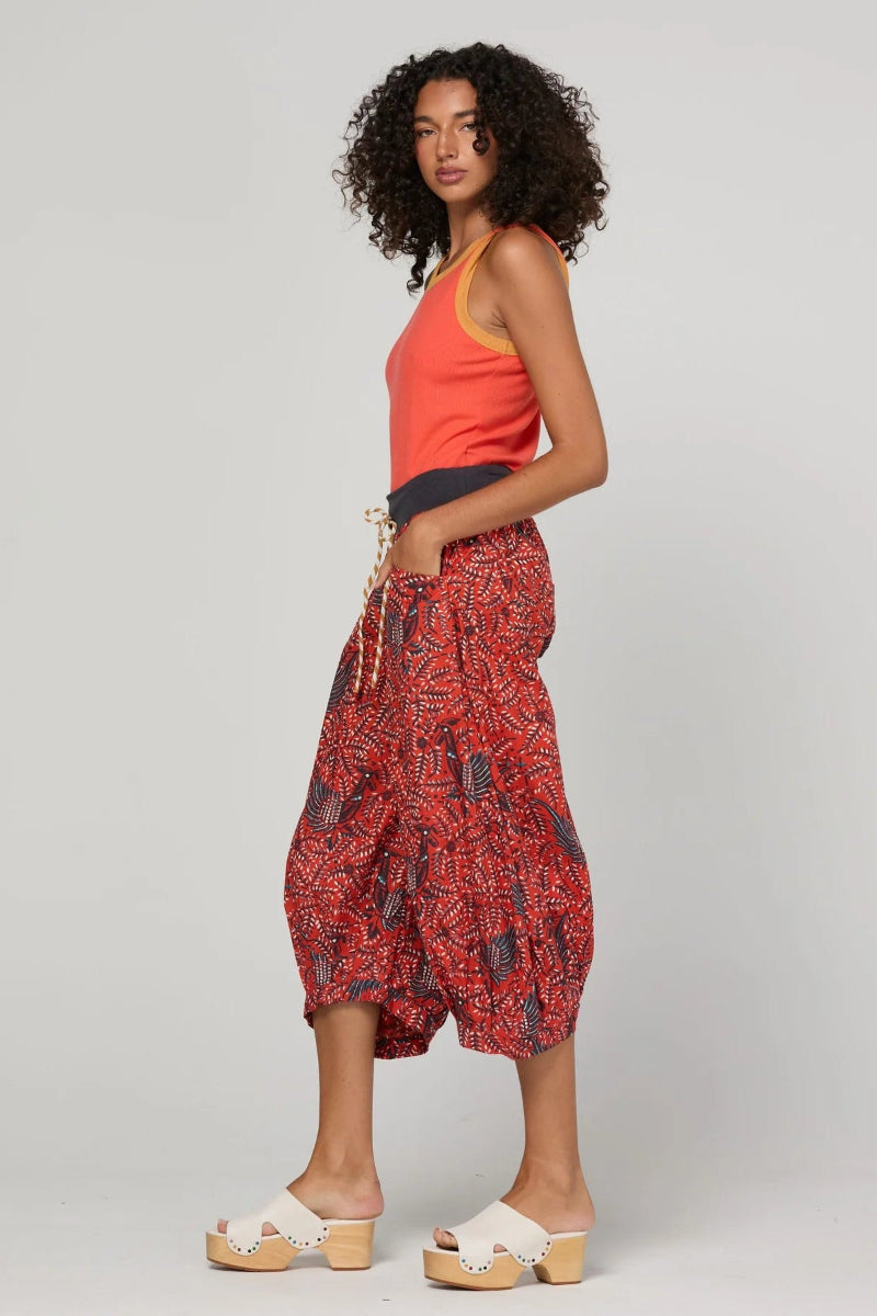 A model wearing a bright orange sleeveless top, red and black patterned harem pants, and white platform slide sandals, standing against a light gray studio background.