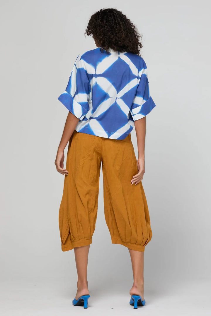 A model is shown from the back wearing a blue and white tie-dye patterned short-sleeve top and mustard yellow cropped pants, paired with blue heels, against a light gray studio background.