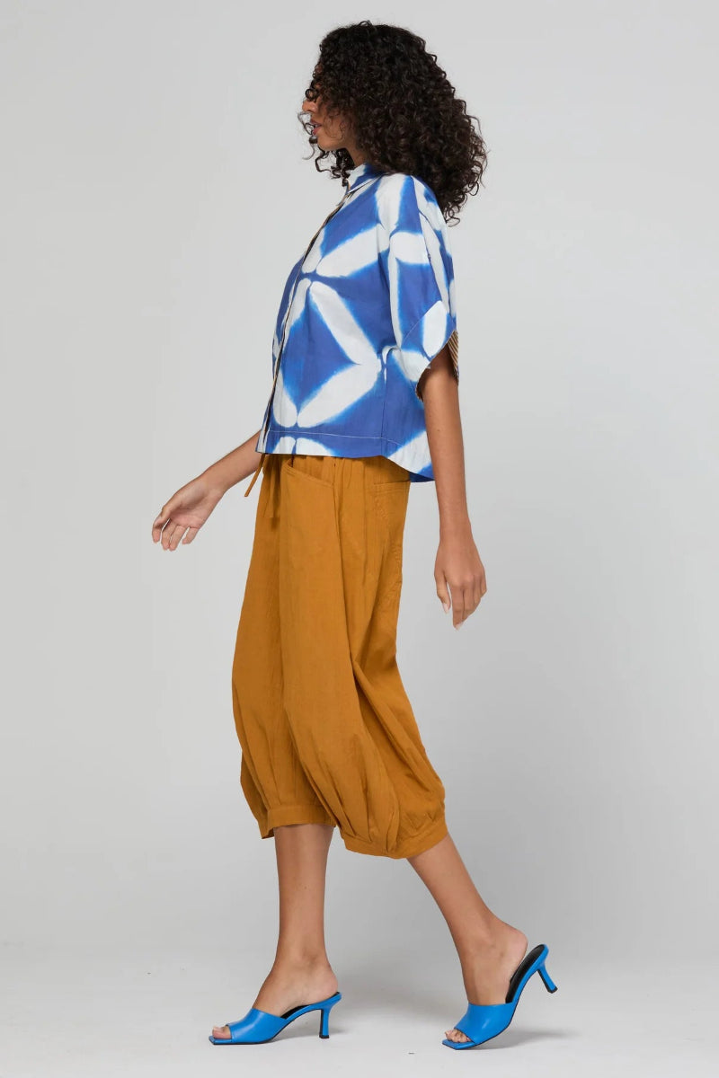 A woman modeling a blue and white geometric patterned short-sleeve top paired with mustard yellow cropped pants and bright blue mule heels, photographed against a light gray studio background.